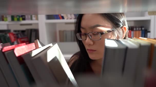 Estudiante asiática estudiando en la biblioteca universitaria. Joven mujer recogiendo libro de estante para el proyecto — Vídeos de Stock