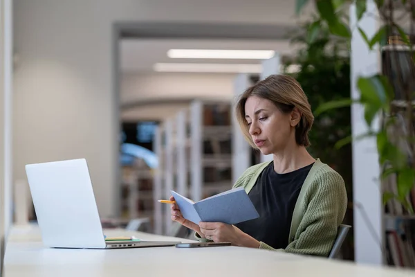 Femme concentrée écrivant la liste des choses à faire dans le carnet tout en travaillant à distance sur un ordinateur portable dans une bibliothèque tranquille confortable — Photo