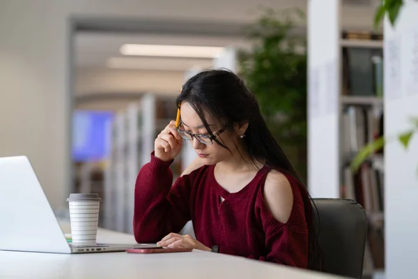 Asiatisk student flicka förbereder sig för examen i biblioteket medan du sitter vid bordet med bärbar dator — Stockfoto