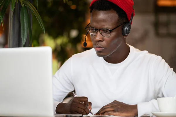 Concentratie zwarte hipster man in hoofdtelefoon, oogkledij studing of werken op afstand op de computer — Stockfoto