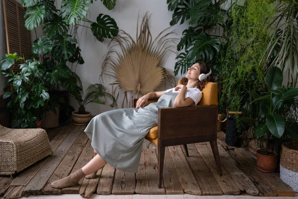 Calma fiorista donna in cuffie moderne ascoltare musica rilassarsi nel giardino di casa con diverse piante da casa — Foto Stock
