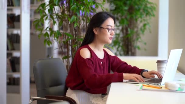 Boldog ázsiai diák lány felkészülni egyetemi vizsgák segítségével laptop könyvtár házi feladatot — Stock videók