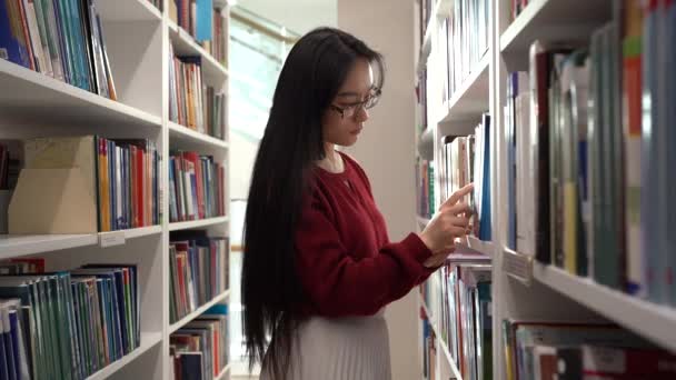 Asijské studentky vyzvednout knihu z police, zatímco stojí mezi knihovnami — Stock video