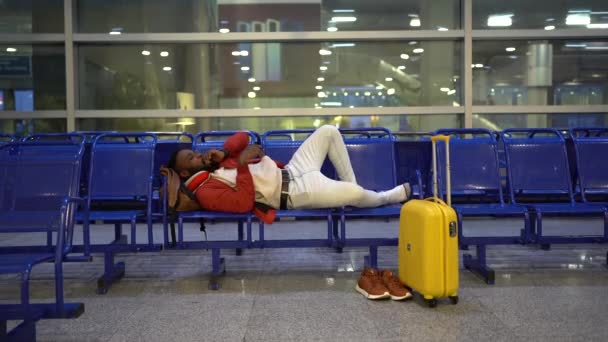 飛行を待っている間にアフリカ人の男が昼寝をする。黒男は空港の荷物の近くのベンチに横たわっていた — ストック動画