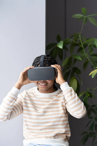 Young amazed excited afro girl wearing VR helmet relaxing with virtual reality on windowsill at home — Stock Photo, Image