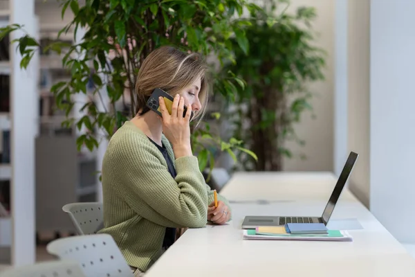 Орієнтована зайнята жінка розмовляє на мобільному телефоні, працюючи дистанційно в читальному залі бібліотеки — стокове фото