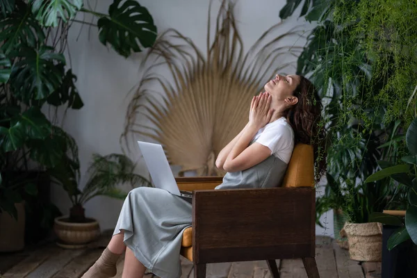 Thoughtful woman with closed eyes sitting on armchair after finishing work on project in homegarden.