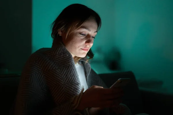 Smutná dívka znuděný rolování přes sociální sítě na mobilním telefonu pozdě v noci v tmavé ložnici. — Stock fotografie