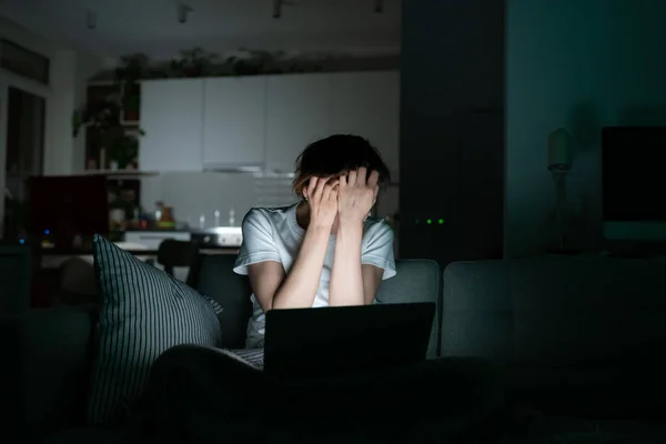 Úzkostlivá žena sedí v noci na gauči a používá notebook, čte si náhlé špatné zprávy a zakrývá obličej dlaněmi — Stock fotografie
