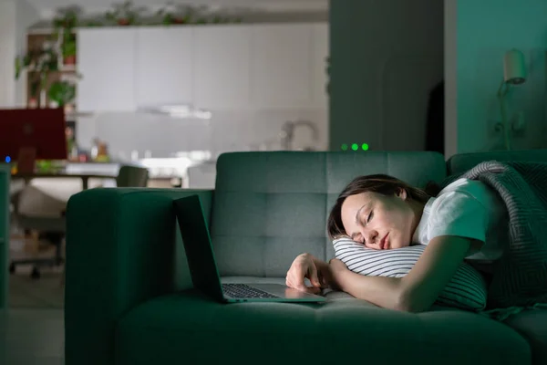 Vyčerpaná mladá žena spí na gauči po práci s notebookem pozdě v noci doma — Stock fotografie