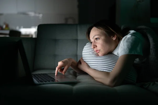 Измученная женщина, лежащая на диване, используя ноутбук поздно ночью дома, не может спать. Бессонница — стоковое фото