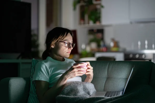 밤에 집에서 노트북 컴퓨터를 사용하여 차나 커피를 마시는 생각깊은 젊은 여자 — 스톡 사진