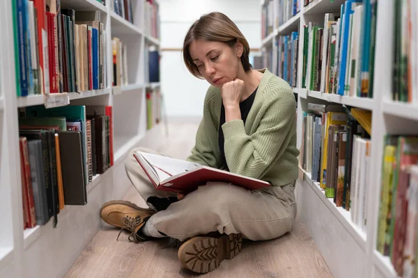 Gericht vrouw van middelbare leeftijd enthousiast kijkend in boek op haar knieën zittend in de bibliotheek op de vloer. — Stockfoto