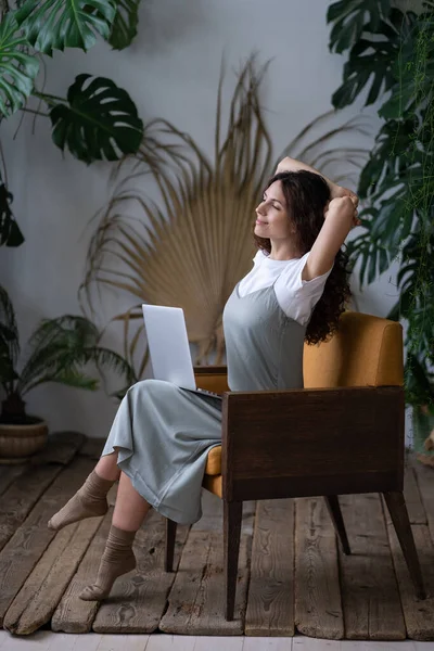 Piękna kobieta z rękami za głową i komputerem na kolanach odpoczywająca, marząca w roślinach otaczających — Zdjęcie stockowe