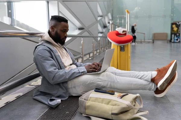 年轻的黑人男子带着手提电脑坐在机场，带着手提箱在网上搜索旅行信息 — 图库照片