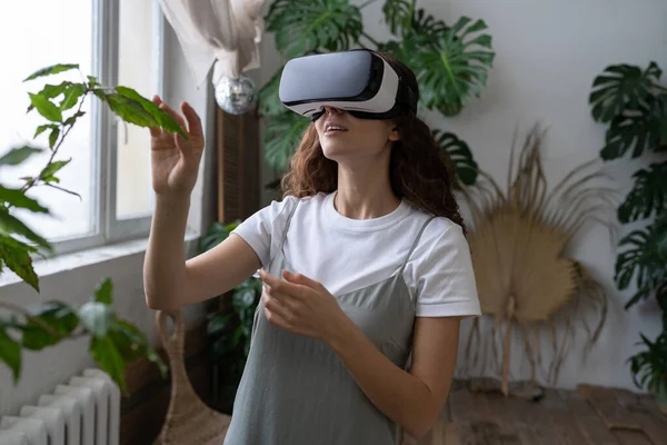 Home gardening, metaverse. Woman using VR glasses in greenhouse, touching plant. Augmented reality. — Stock Photo, Image