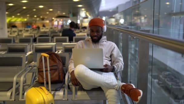 Uśmiechnięty hipster czarny człowiek siedzi w terminalu lotniska za pomocą laptopa nosić słuchawki oglądać film — Wideo stockowe