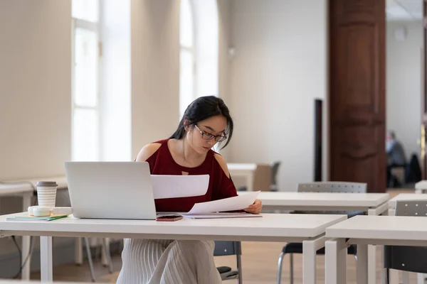 Asiatisk flicka college student i glasögon läsa material samtidigt förbereda sig för test eller examen i biblioteket — Stockfoto