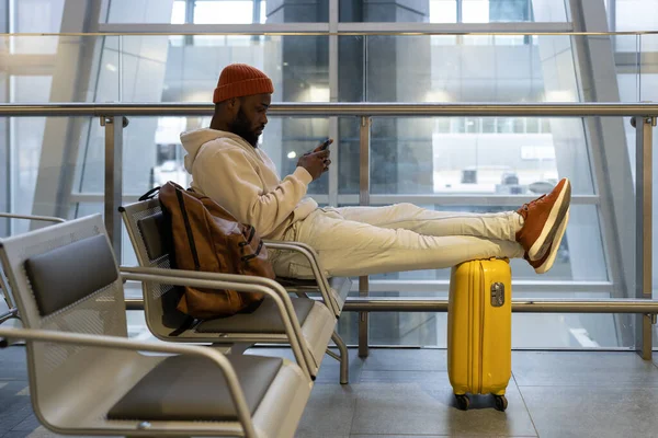 Thoughtful young hipster black man sitting in airport terminal using smartphone and waiting landing