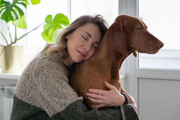 Middle age woman hugging her beloved Wirehaired Vizsla dog at home. Female owner kissing and embracing her lovely pet in living room. Animal therapy, stress relief concept.
