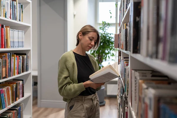 Pensive woman university professor reading textbook standing near bookshelf. Concentrated female mature student spending leisure time in library while preparing for test exam. Second higher education.