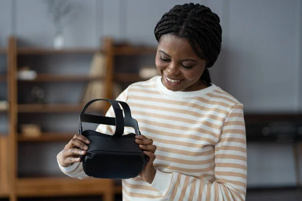 Curious black woman holding futuristic innovative VR glasses in hands at home. Young african american lady testing modern virtual reality headset for video games. Future technology, augmented reality
