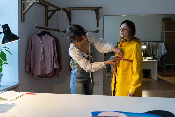 Making measures before tailoring. Fashion designer workshop with female tailor measuring and fitting bespoke rain jacket. Young woman small dressmaking business owner at workplace. Needlework concept