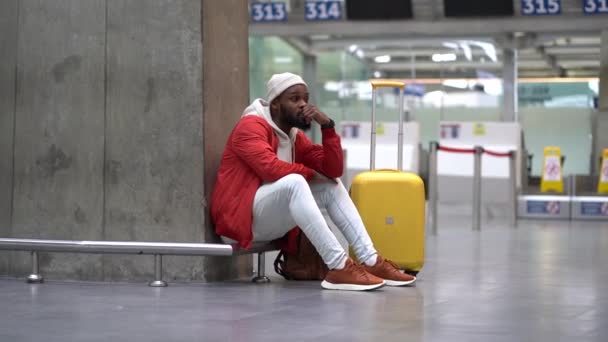 アフリカ系アメリカ人の男性が空港で動揺し 彼のフライトは遅れています 緊張した旅行者の男性の飛行機の手荷物を空のターミナルに座って待っている 空港での長い夜の接続の疲れ男 — ストック動画