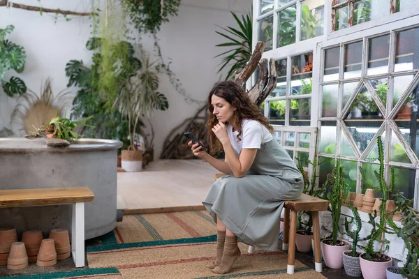 Serious young woman owner holding smartphone reading media news, sitting in home garden. Thoughtful female gardener using phone, thinks over received notification internet information in plant store.