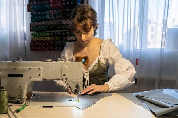 Clothing production business: young studio owner female at workplace sew clothes on modern sewing machine in professional atelier. Tailoring hobby, garment industry and needlework occupation concept