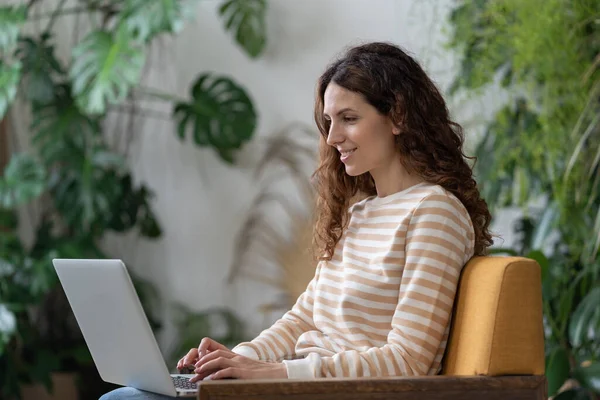 Joyful woman sitting on armchair relaxing while browsing online shopping website. Happy girl surfing net, chatting with friends during free time at cafe. Young female doing research work for business.