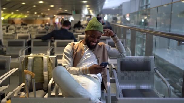 携帯電話を使用しながらラウンジ空港の場所に座って白いジーンズの幸せな若いアフリカ系アメリカ人の旅行男 ヒップスターで緑の帽子ビーニー腕時計楽しいビデオオンラインで携帯電話 サーフィンソーシャルメディア — ストック動画