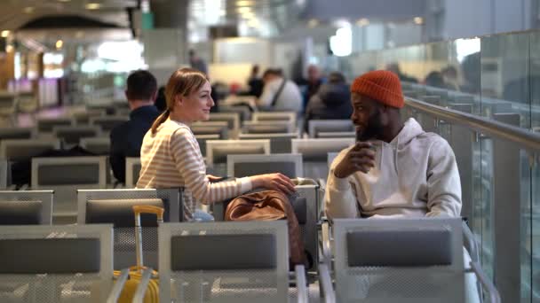 飛行遅延中の新しい知人 アフリカ系アメリカ人のヒップスター男性がコーヒーを飲み かなり白人女性の旅行者がターミナルで椅子に座っているさまざまなトピックについて話しています 旅行の概念 — ストック動画