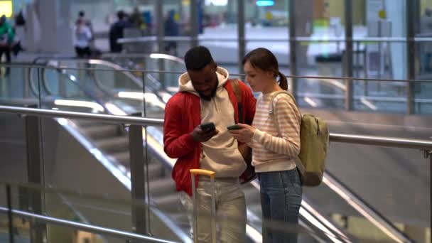 若い異人種間の旅行カップルは 空港で一緒に立っている間 スマートフォンを使用します 多民族の友人アフリカ人男性と原因アジアの女性は フライトをチェックしたり 携帯電話経由でオンラインチェックインを行います — ストック動画