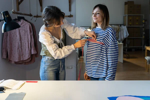 Fashion designer woman or seamstress takes measurements with measuring tape of girl client before tailoring clothes to order. Young female small dressmaking business owner at workplace. Small business
