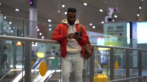 若いですアフリカ人男性旅行者スーツケースとともに携帯電話で話す駅 幸せな黒旅行男作る電話コールしながら待っていますのために友人で現代の空港ターミナル — ストック動画