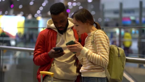 多人種間の旅行者のカップルは 空港ターミナルに立って フライトまたはオンラインチェックインをチェックするためにスマートフォンを使用します モバイルアプリでチケットを探している若い黒人男性と白人女性 一緒に旅行する — ストック動画