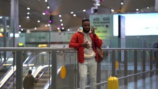 若いアフリカ系アメリカ人の魅力的な男性が空港や鉄道駅でスマートフォンで話すスーツケースとエスカレーターの近くに立っています 男は旅行パートナーと接続するためにダイヤルの携帯電話番号の周りに見える — ストック動画