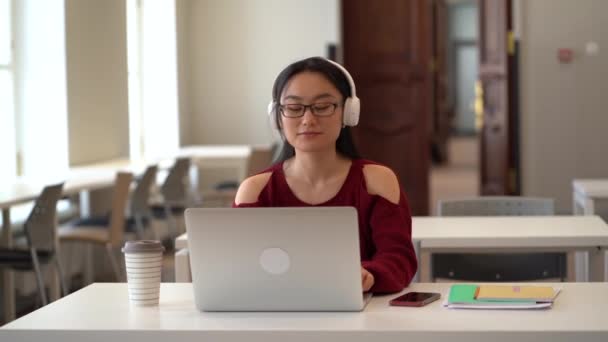 Een Glimlachende Aziatische Vrouw Met Een Draadloze Hoofdtelefoon Typt Een — Stockvideo