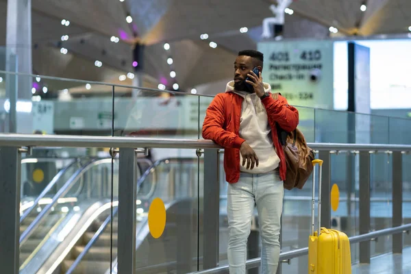 Young African American man traveler talking on mobile phone, making call while standing with suitcase at airport or railway station, black guy talking with family or friends after arriving in terminal