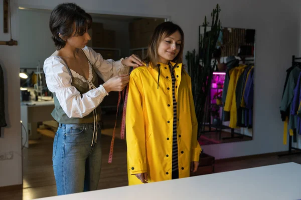 Making measures before tailoring. Fashion designer workshop with female tailor measuring and fitting bespoke rain jacket. Young woman small dressmaking business owner at workplace. Needlework concept