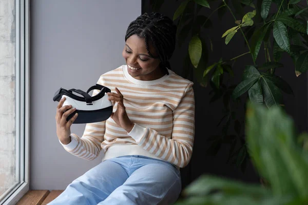 Young black woman testing virtual reality glasses, sitting on windowsill and looking curiously at VR headset in hands. Joyful african lady enjoying first augmented reality experience at home