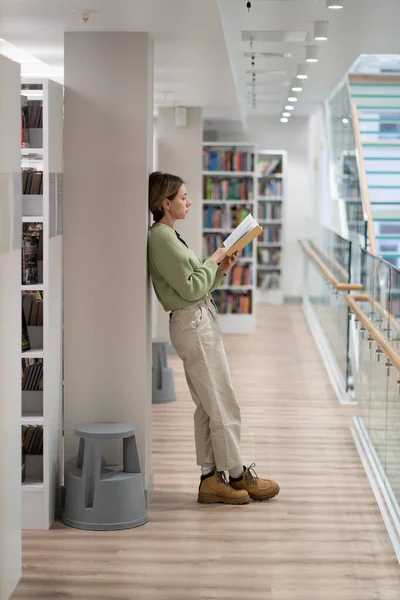 Full length of pensive middle-aged woman book nerd reads interesting paper covered book in library, focused pensive female devoted to reading and study, mature student with textbook near bookcase