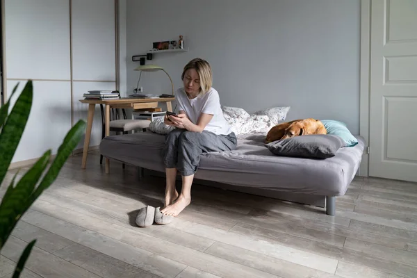 Woman sitting on sofa with sleepy dog using smartphone in hands wait for boyfriend call or text feels depressed. Thoughtful female think about relationships problems. Mental health, loneliness concept