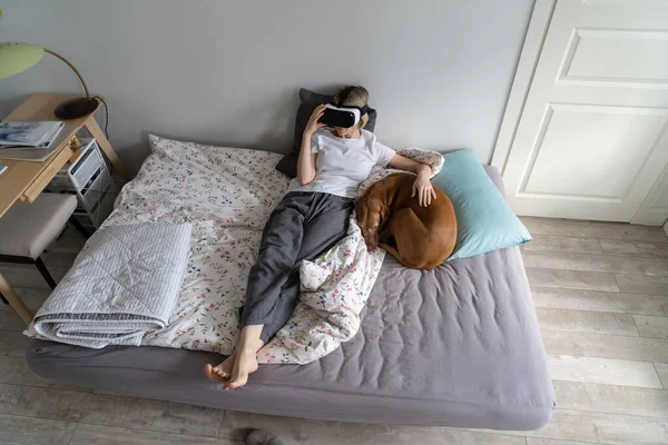 Virtual entertainment at home. Calm young female in virtual reality goggles resting at in relaxing virtual environment at home with lovely dog. Woman wearing VR helmet, using futuristic technology