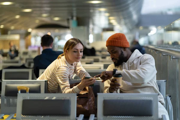 Attractive European woman asks African American hipster man to help connect wifi. New acquaintances during flight delay. Traveler checking boarding time with smartphone app. Travel interaction concept