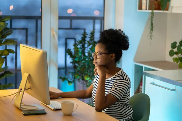 Confident African American woman looking at computer screen, thoughtful graphic designer pondering idea, strategy, working on research project in kitchen at home at night. Distance education.