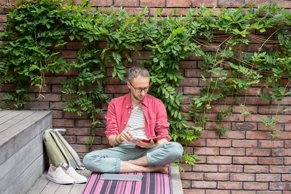Enthusiastic man sitting cross-legged on wooden stairs near green plant wall, reading favourite book, searches for information and conduct research without internet, getting second higher education