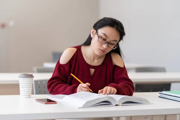Smart asian girl reading textbook and taking notes writing outline while preparing for exam, sitting in empty quiet university reading room or library, korean female student learning in classroom