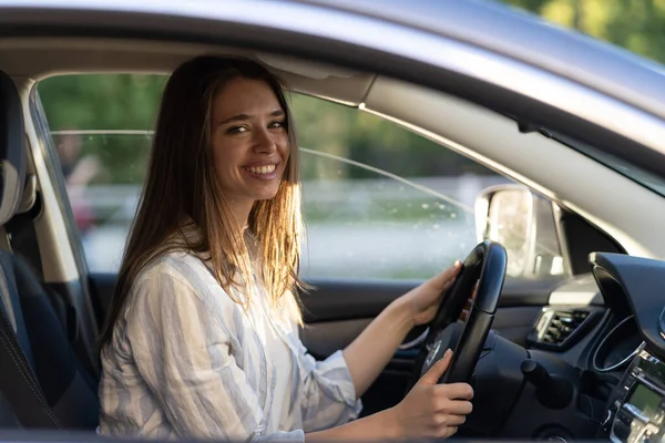 Happy young girl sitting on driver seat in new car joyful smiling hold hands on wheel. Cheerful female driving vehicle looking through open window. Successful woman car owner or getting driver license